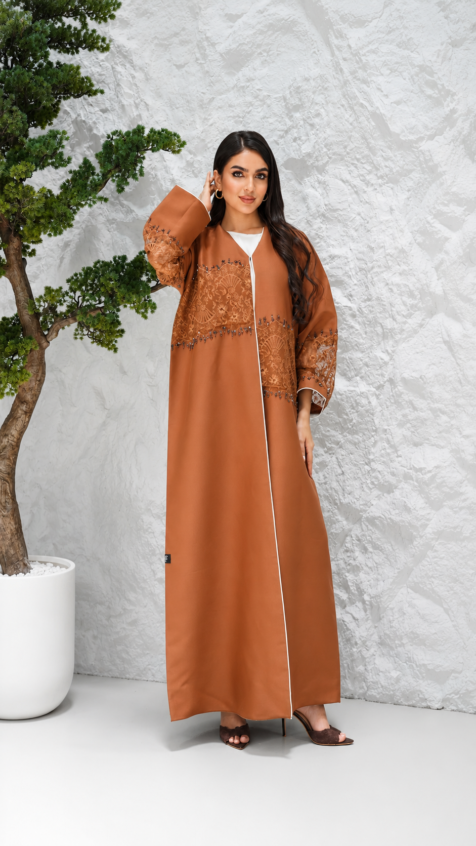 A woman models a long, rust-brown abaya dress with intricate embroidery on the chest and sleeves, styled over a white inner top. She is standing in front of a textured light gray wall next to a potted bonsai tree.
